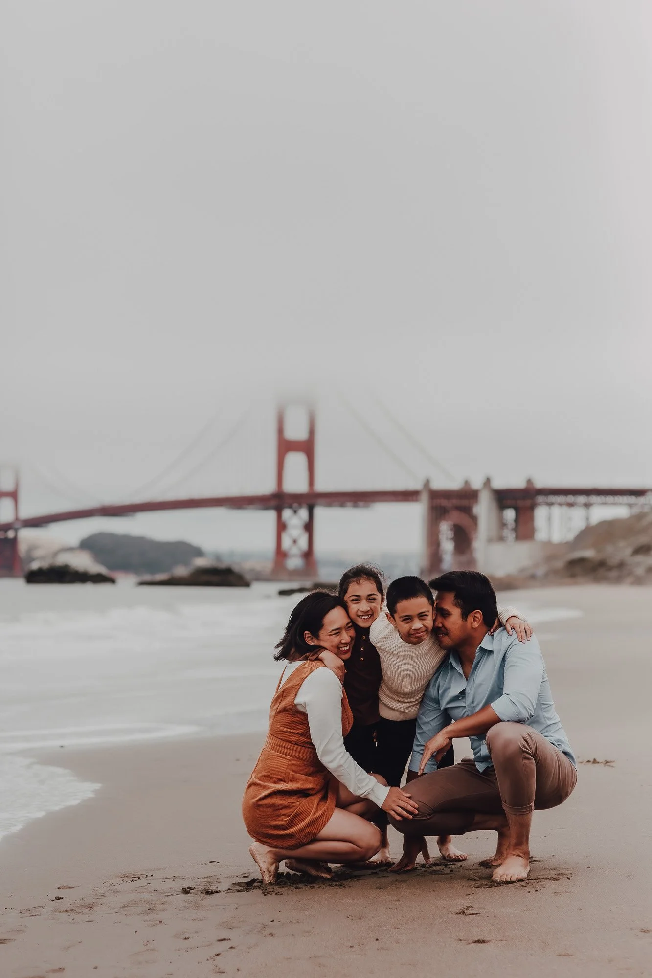 Family of four smiling and hugging on a beach with the Golden Gate Bridge in the background.