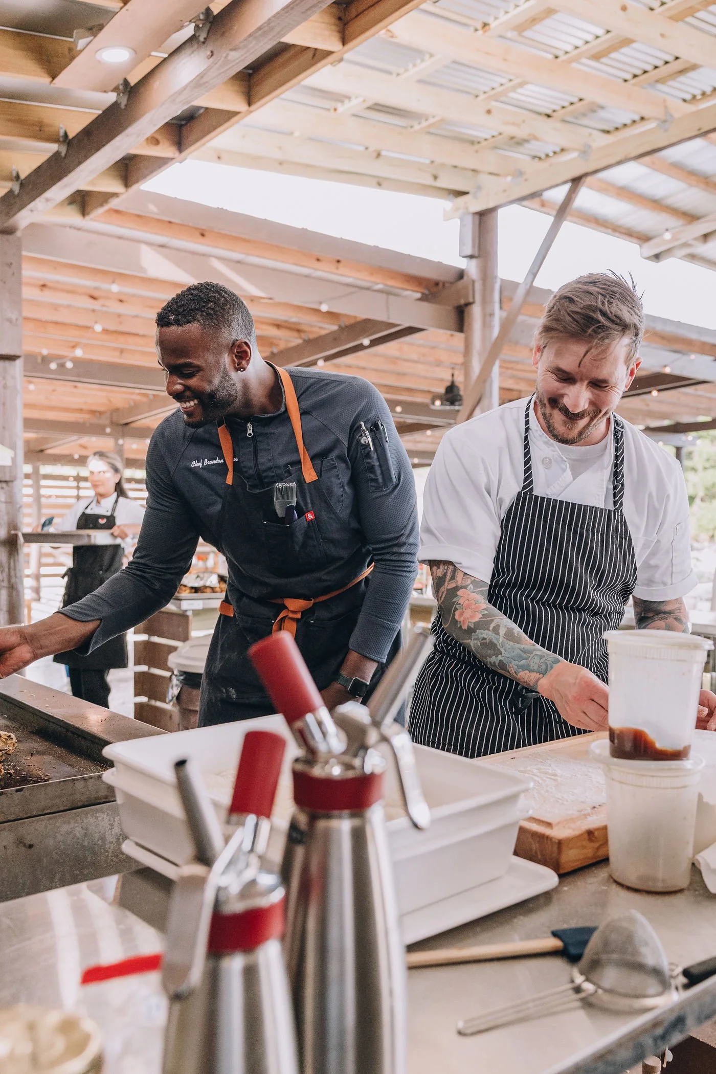 Two male chefs smiling and preparing food in an open-concept kitchen with wooden beams and sunlight.