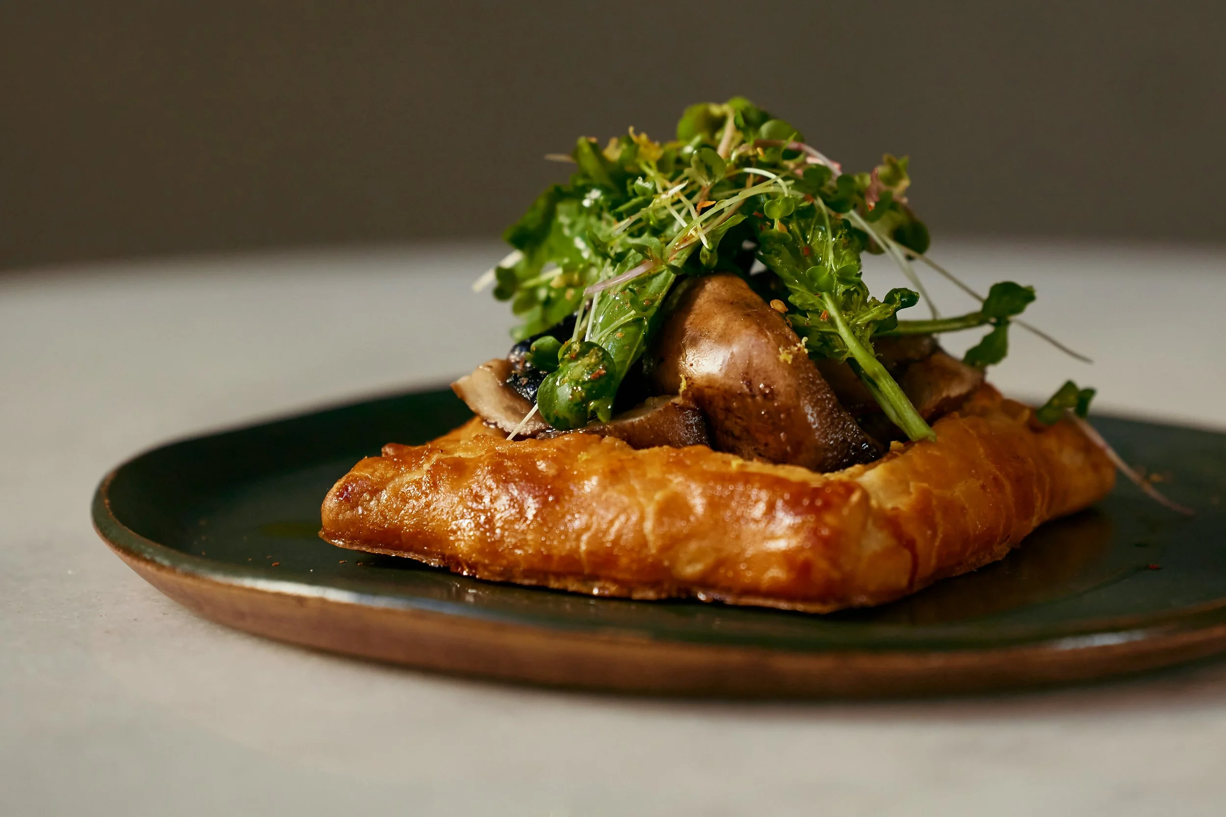 A gourmet dish featuring a crispy fried pastry shell filled with sautéed mushrooms and topped with fresh microgreens on a dark ceramic plate.