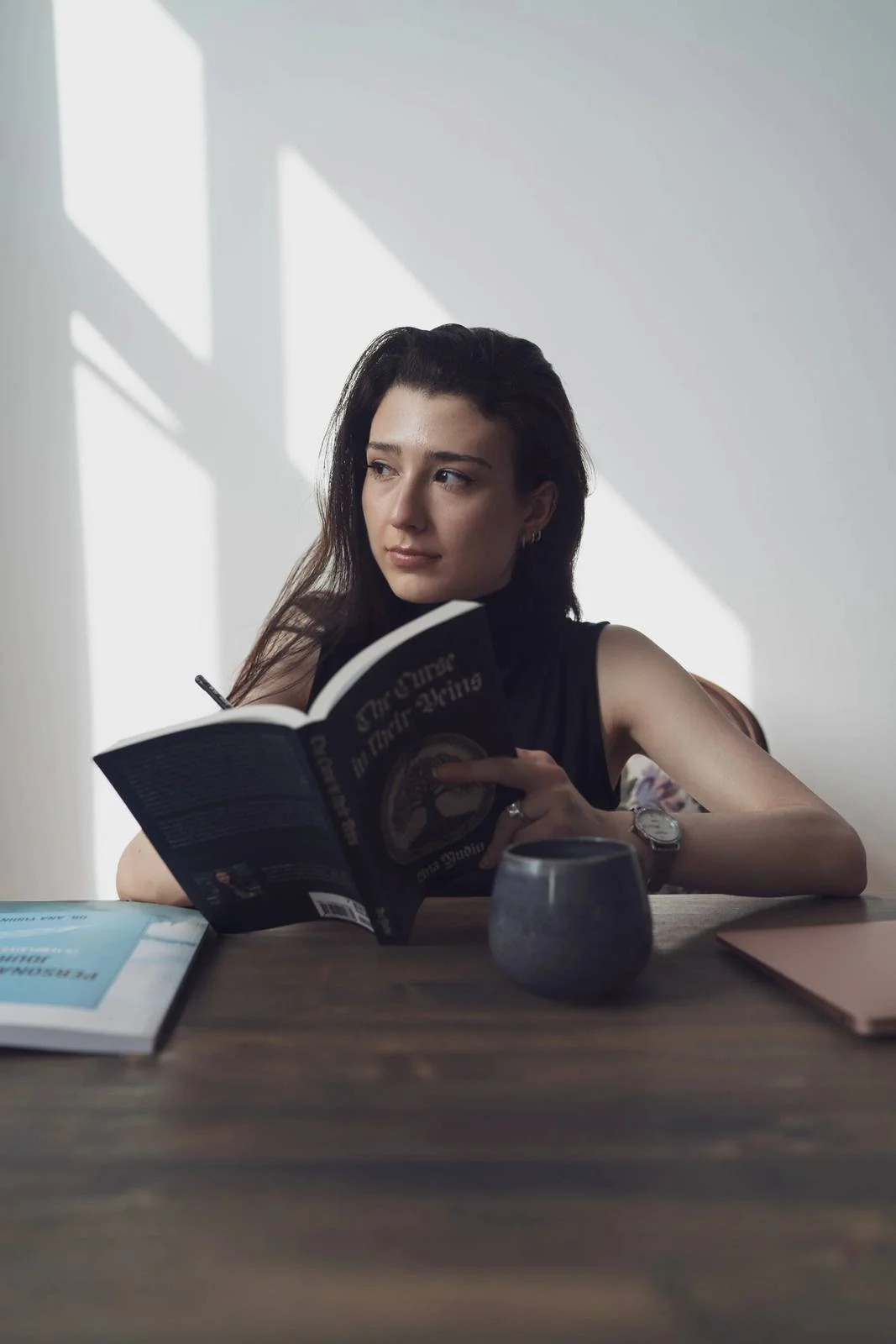 A young woman with dark hair and earrings sits at a desk, reading a book titled "The Power of Full Engagement" with sunlight casting shadows on the wall behind her.