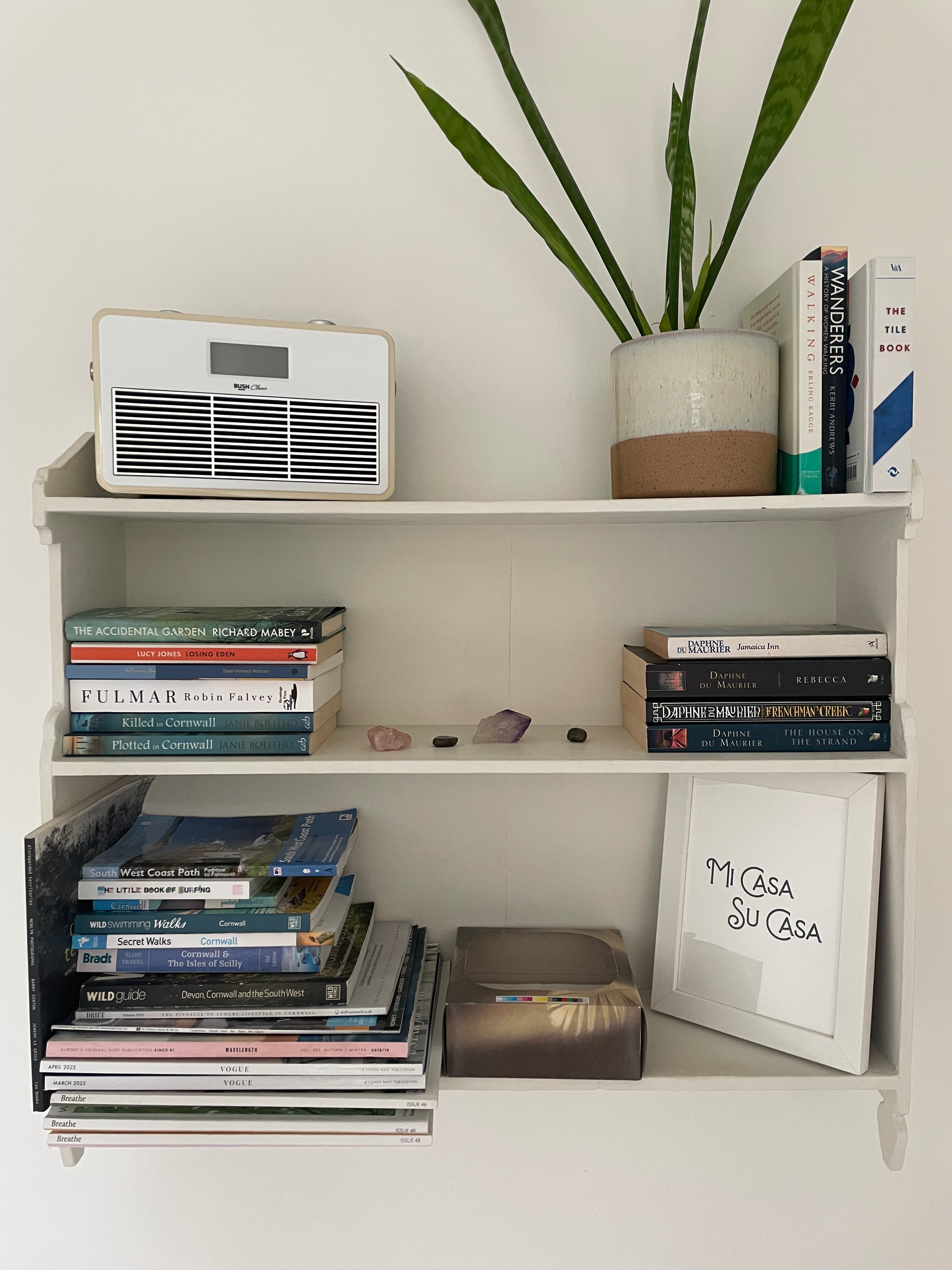 A white bookshelf holding books, a potted plant, a silver box, a framed sign that reads 'MI CASA SU CASA,' a white fan, and decorative rocks.