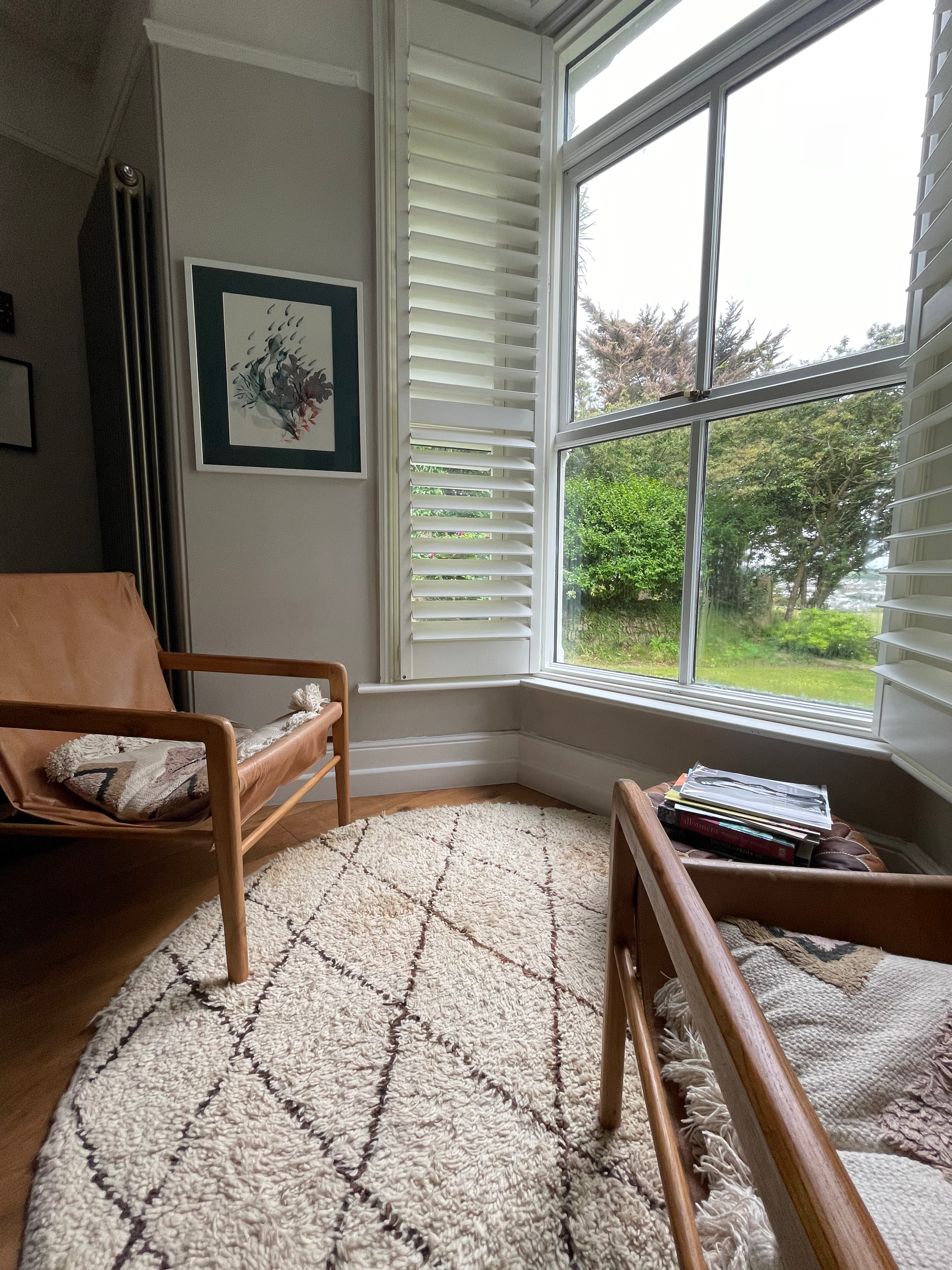 A cozy living room corner with large window, white blinds, wooden chair with throw pillow, bookstand, and a framed abstract art piece on the wall, with a view of green trees outside.