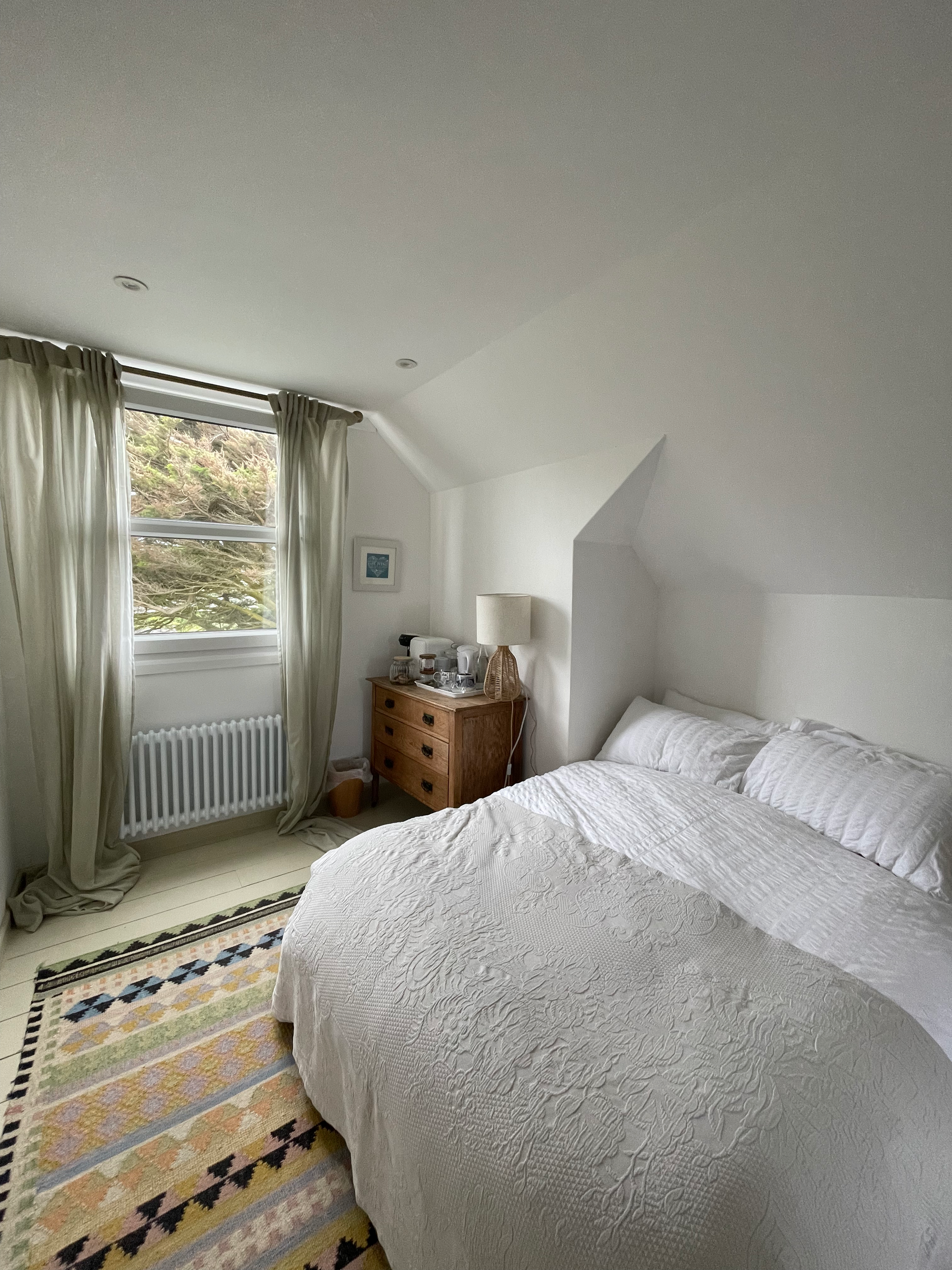 A bedroom with a white quilted bedspread, a wooden nightstand with a lamp, and a window with cream curtains, a colorful rug on the floor.