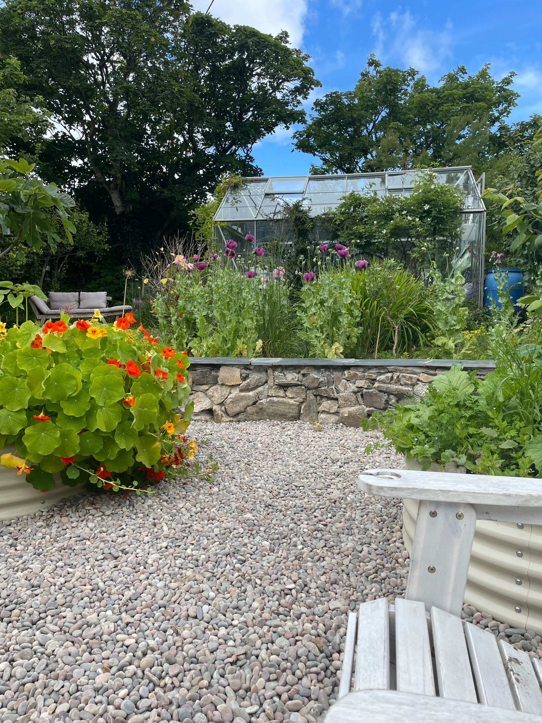 A garden with a greenhouse, colorful flowers, surrounding trees, gravel path, and outdoor furniture under a blue sky.