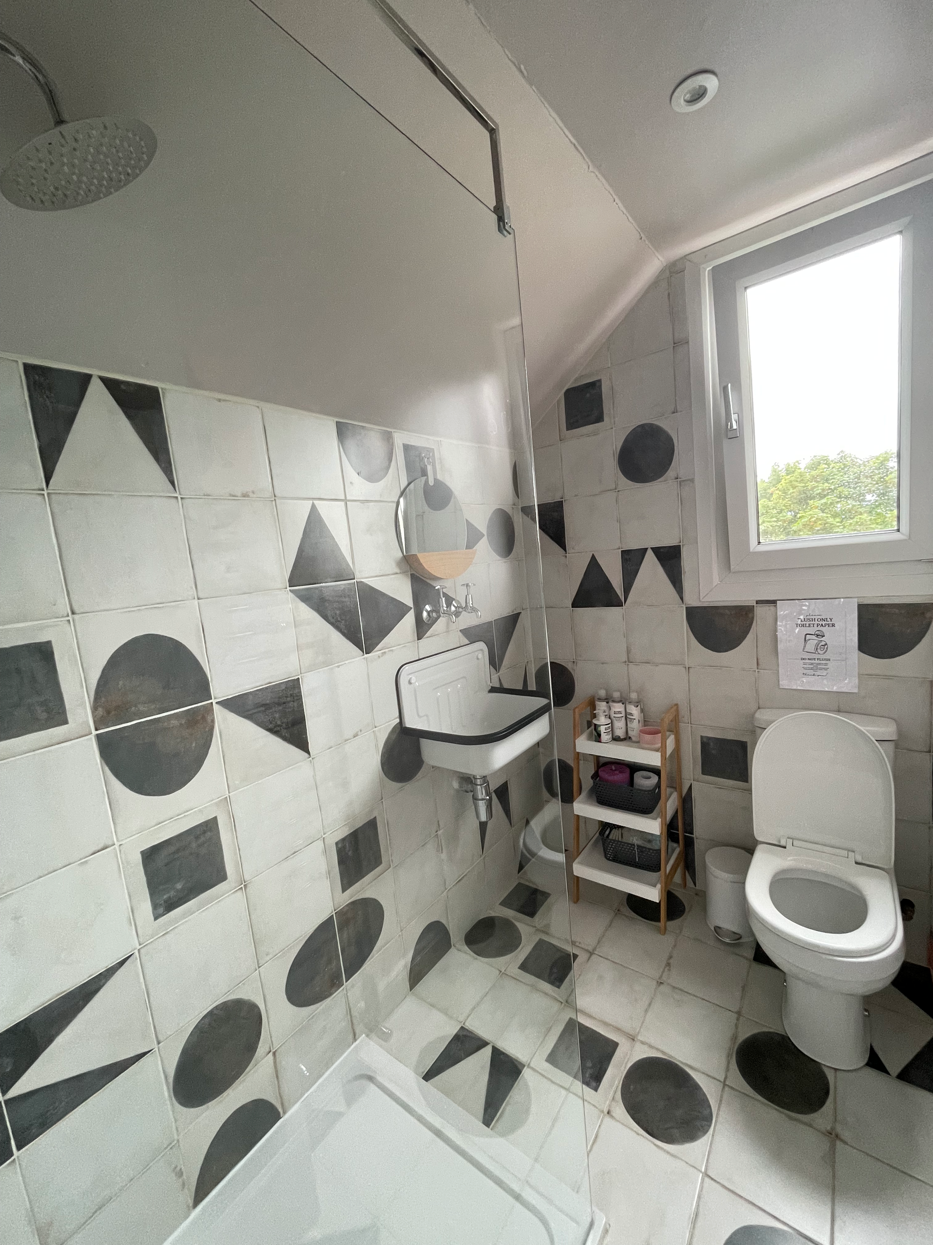 A bathroom with a window showing green trees outside. It has black and white geometric patterned tile floors and walls. There is a white toilet, a small white sink, and a clear glass shower enclosure. A wooden shelf holds toiletries, and a mirror is 