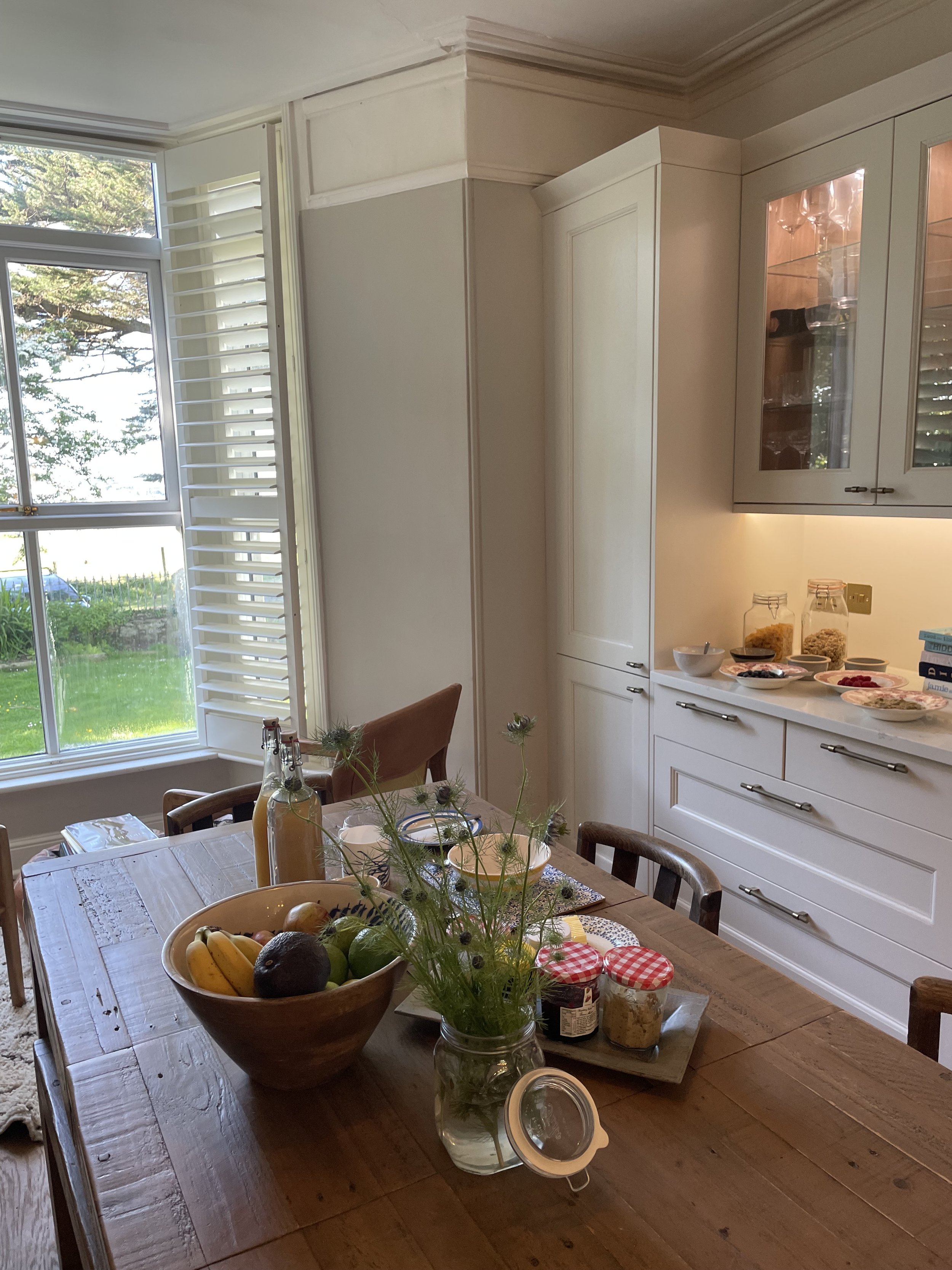 A dining table with a bowl of bananas, an avocado, limes, and other fruit, surrounded by wooden chairs, set with plates and bowls, in a bright kitchen with large windows, white cabinets, and trays of breakfast foods on the counter.