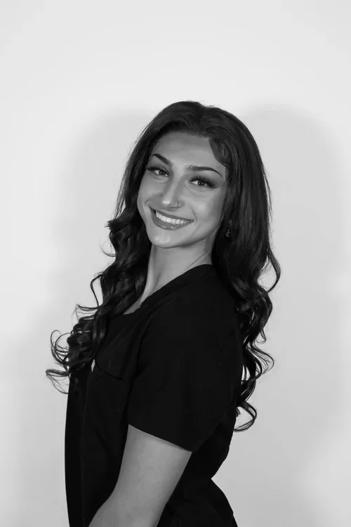A woman with long wavy dark hair, smiling, wearing a black top, standing in front of a plain light background.