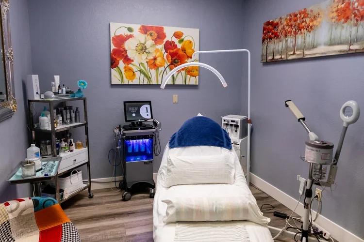 Medical treatment room with a bed, medical equipment, shelves with supplies, floral artwork, and a monitor.