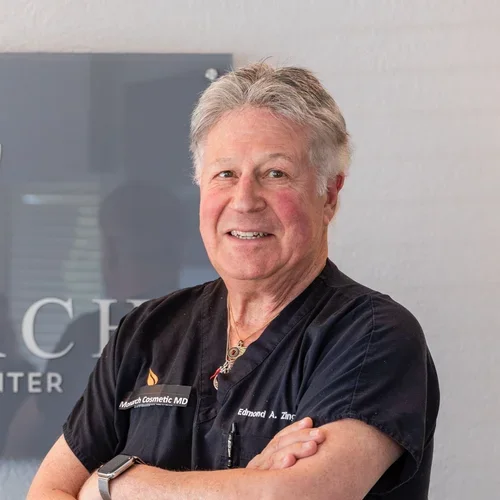 A smiling male doctor wearing a black scrubs uniform with a name tag that reads 'Edmond A. Zinc' and 'Monarch Cosmetic MD', standing with arms crossed in a medical office.
