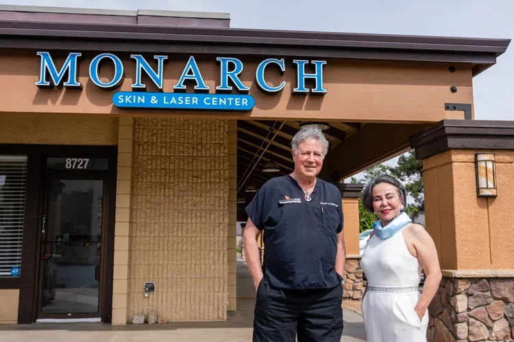 A man and woman standing outside Monarch Skin & Laser Center. The man is wearing a dark medical scrub, and the woman is in a white dress with her hands in her pockets. The building has a brown and beige exterior with a sign that says 'MONARCH Skin & Laser Center' above them.
