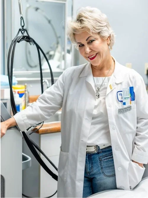 A smiling senior female doctor with short gray hair, wearing a white lab coat, standing in a medical or laboratory setting.