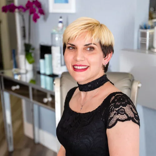 A woman with short blonde and dark hair, wearing a black lace top and choker, smiling in an indoor setting.