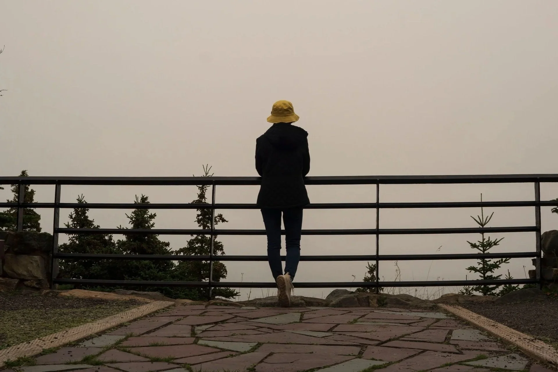 A person in a bucket hat standing at a fence that is at the edge of a foggy ocean overlook