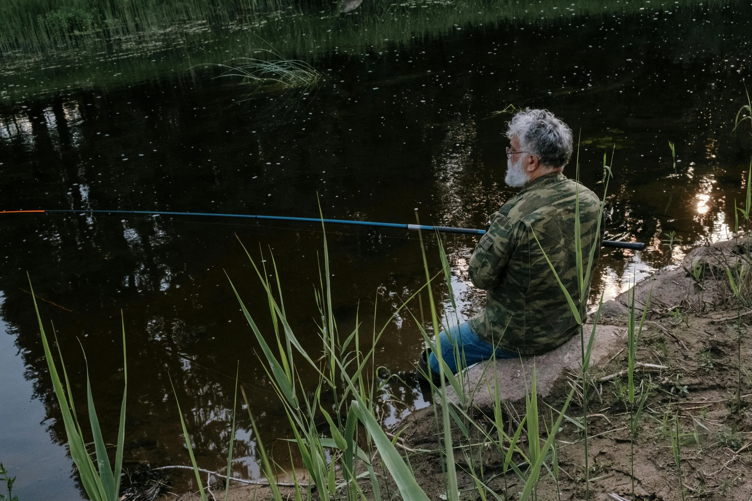 An older man fishing on the bank of a pond or river.