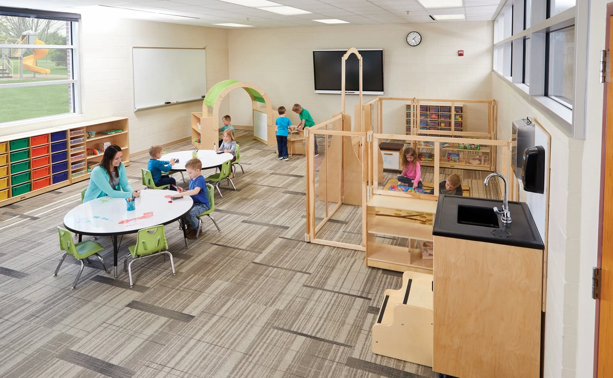 A bright PreK and early learning classroom layout by Oxilium, featuring built-in storage cubbies, colorful floor dot soft seating cushions, a circular group table, and a half-moon crescent desk integrated with interactive edtech for discovery-based p