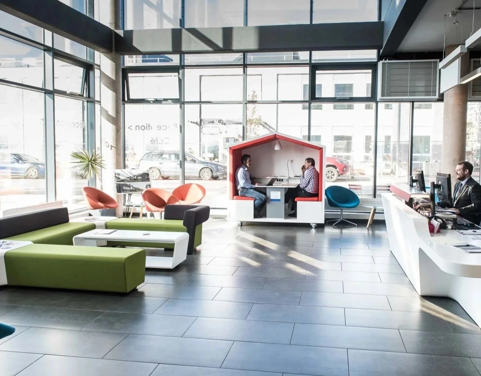 Modern office lobby featuring large glass windows, colorful flexible soft seating, an inclusive NOOK huddle pod, and a dedicated workspace with integrated AV technology solutions designed to support neurodivergent minds.