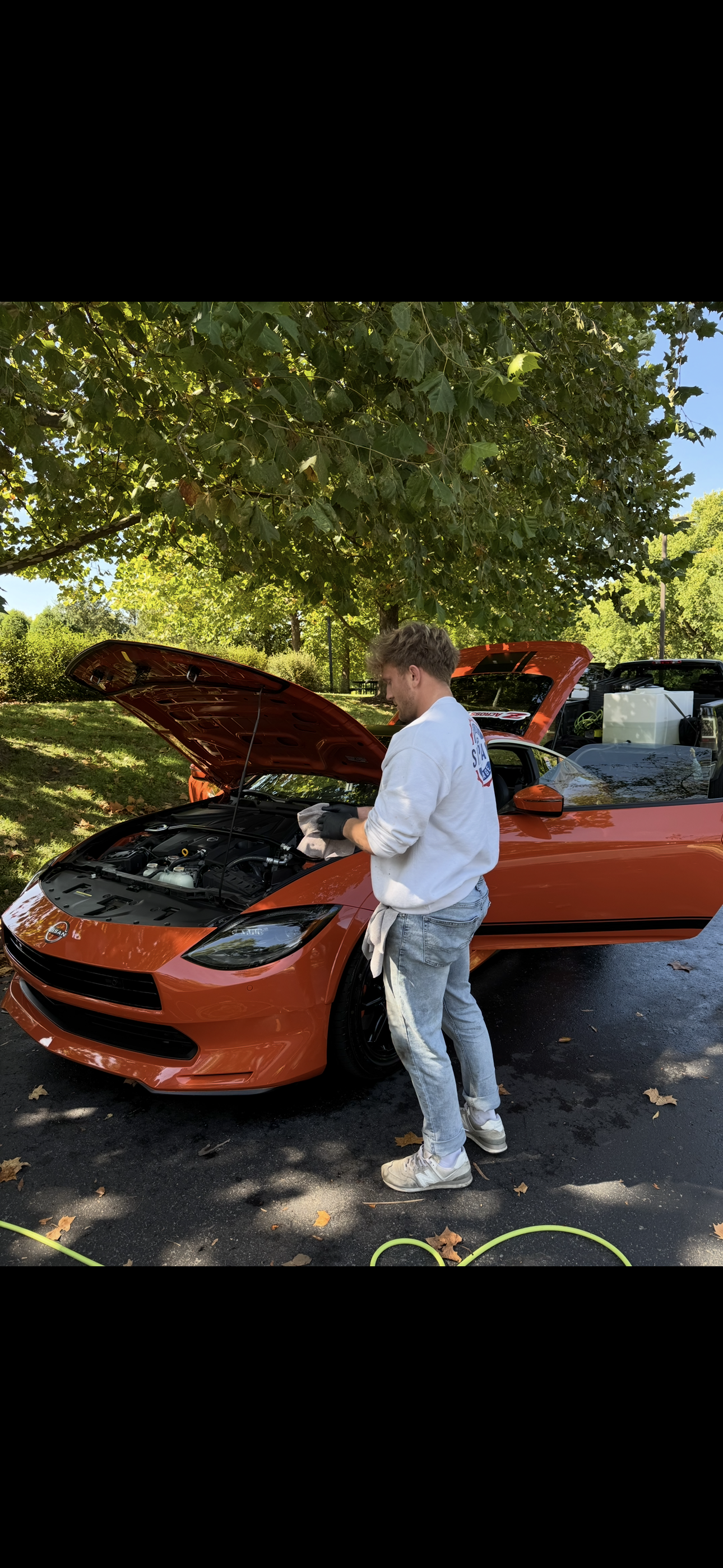 A man cleaning a wire-spoked wheel of a vintage car with a rag. He is bent over, wearing a white t-shirt, light-colored jeans, white sneakers, and has towels tucked into his back pocket. There is cleaning equipment nearby, and the car is dark-colored.