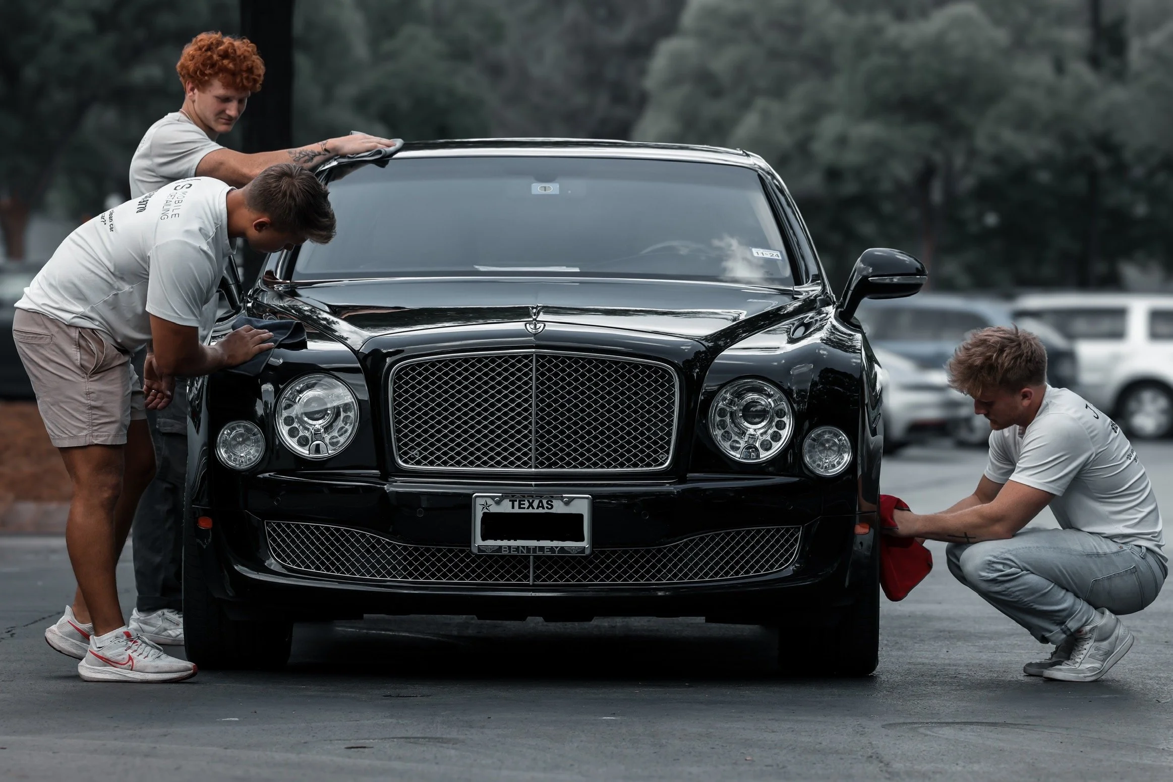 Three young men cleaning and polishing a black luxury car in a parking lot.