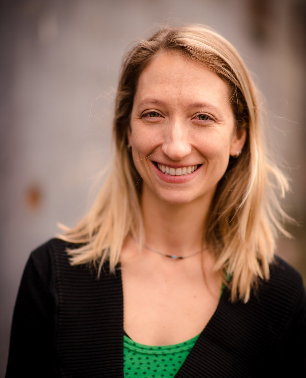 Close-up of a woman with blonde hair smiling outdoors, wearing a black jacket and a green top.