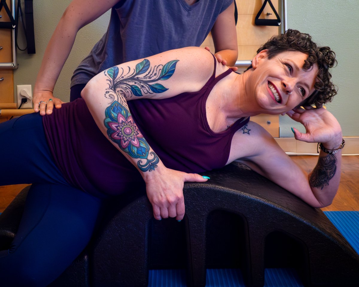 A woman with curly hair and tattoos smiling while doing a side stretch exercise on a Pilates arc, assisted by a pilates trainer.