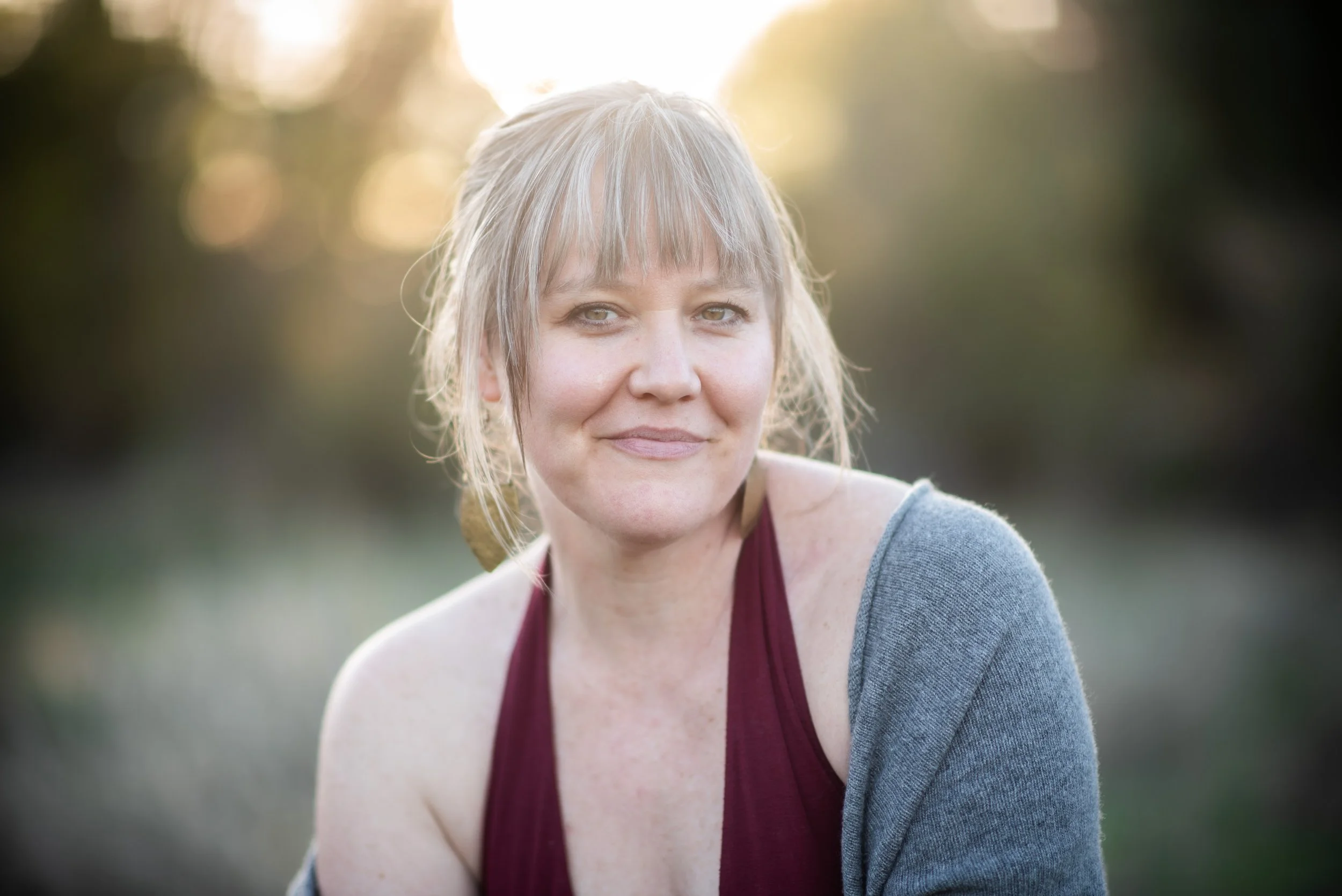 A woman with blonde hair and light skin outdoors with blurred trees in the background, wearing a gray cardigan and a maroon top.