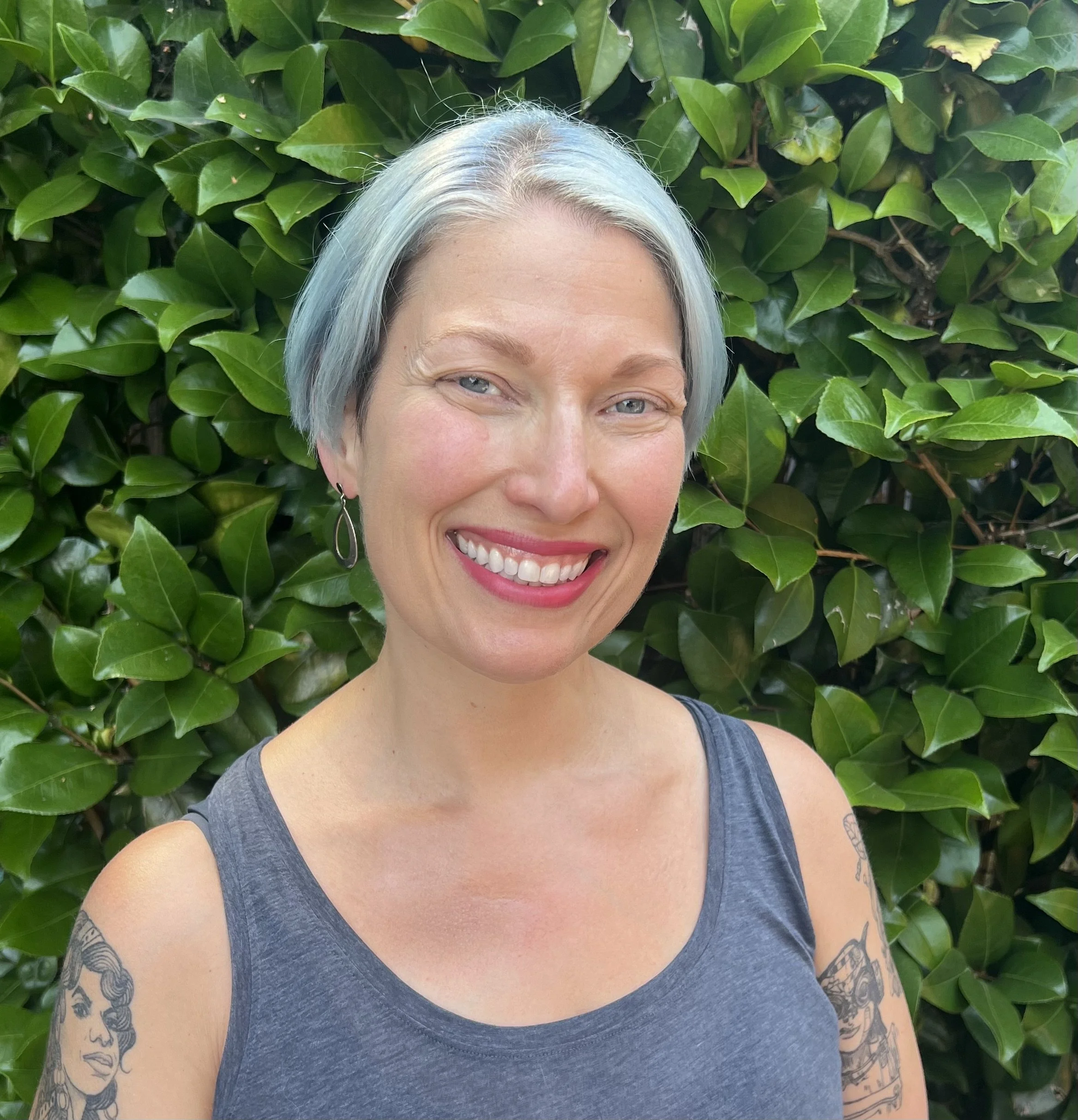 A woman with short silver hair, light blue eyes, and a big smile showing her teeth, taking a selfie outdoors. She is wearing a dark gray sleeveless top and has visible tattoos on her arms. The background is filled with green, shiny leaves.