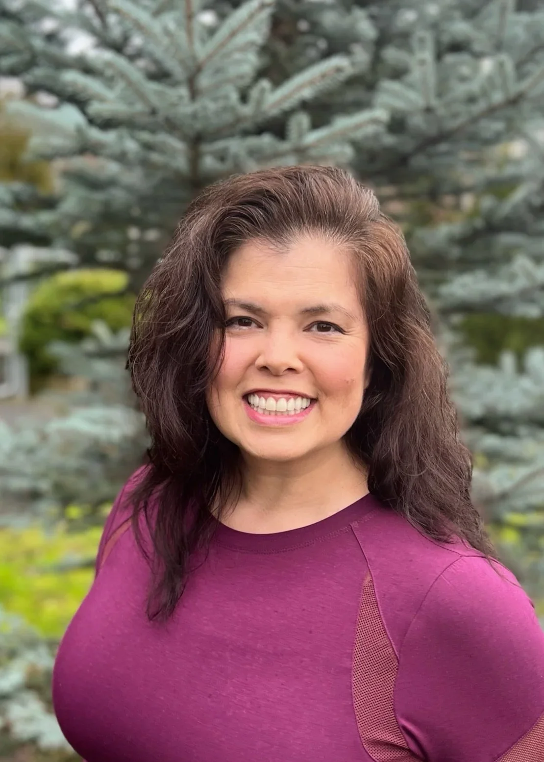 Smiling woman with wavy brown hair wearing a purple shirt standing outdoors in front of a large blue spruce tree.
