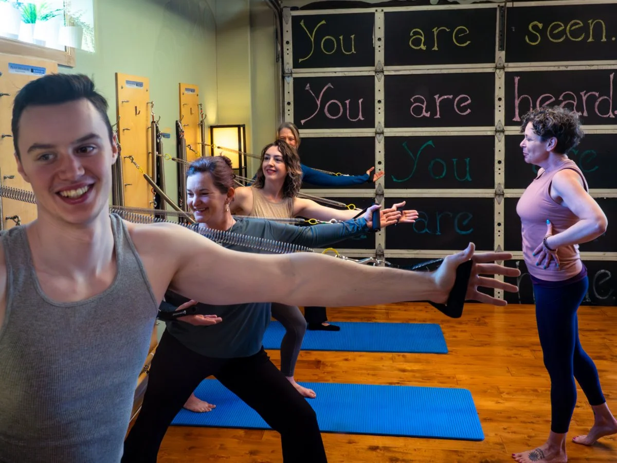 A group of people participating in a yoga class in a studio with a motivational wall that says 'You are seen. You are heard.' The instructor is at the front, and the participants are extending their arms forward in a yoga pose.