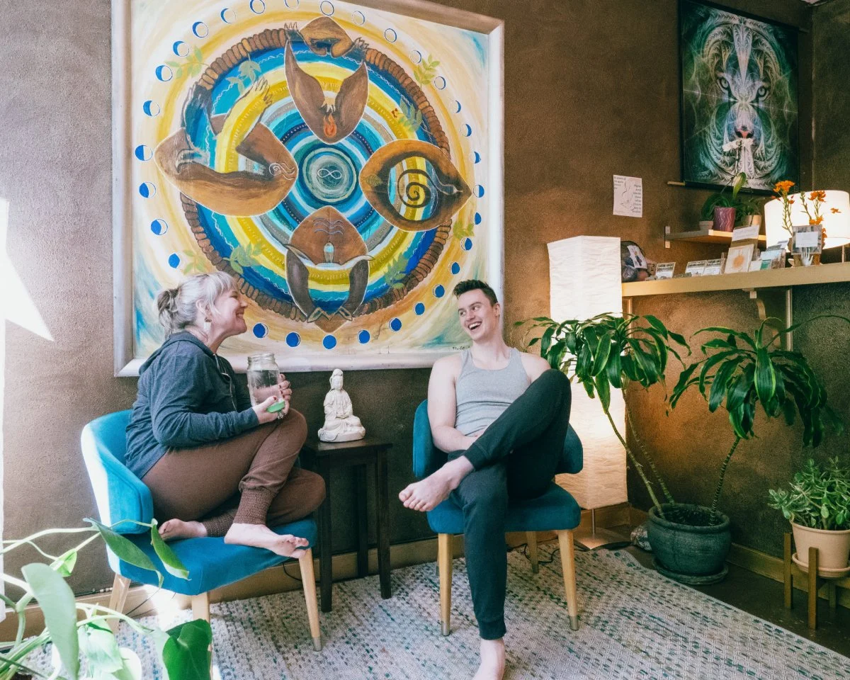 Two people, a woman and a man, are sitting and laughing in a cozy room with vibrant artwork. The woman is holding a jar and sitting cross-legged on a blue chair, while the man is sitting on an adjacent blue chair with one leg crossed over the other. There are large potted plants, a lamp, and decorative items on shelves, with colorful paintings on the wall.