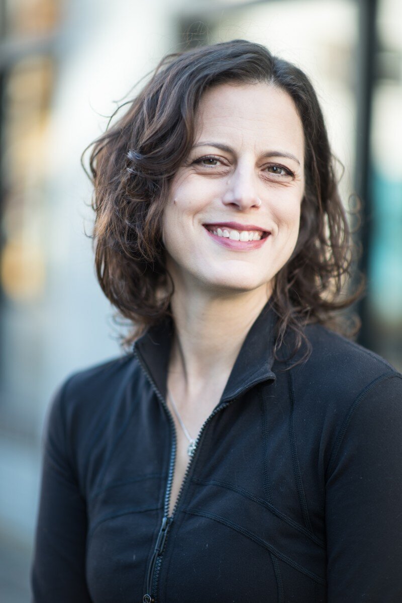 A woman with shoulder-length curly brown hair smiling outside, wearing a black zip-up jacket.