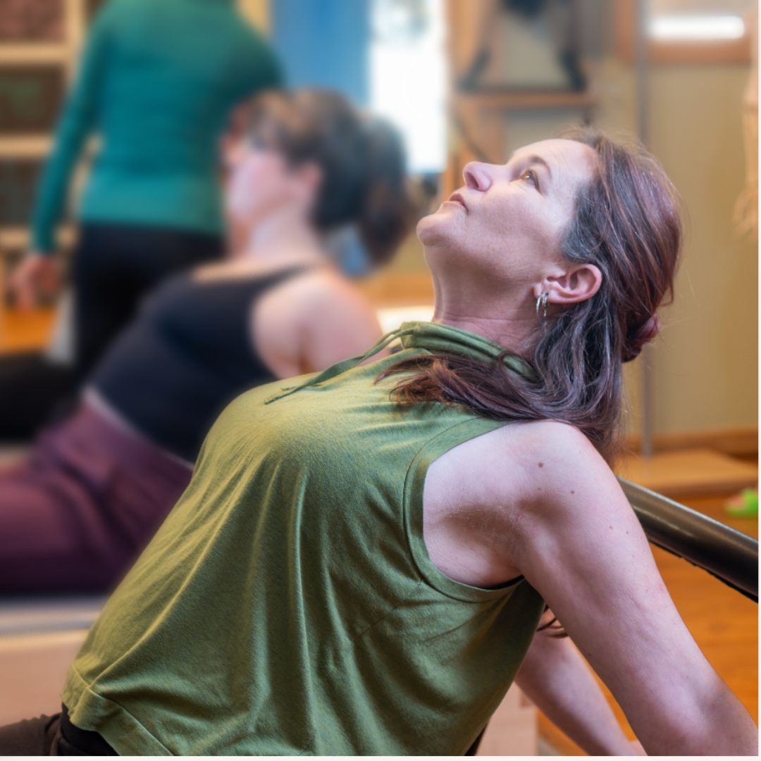 A woman with long brown hair and earrings leaning back and looking up during a workout in a gym, with other women exercising in the background.