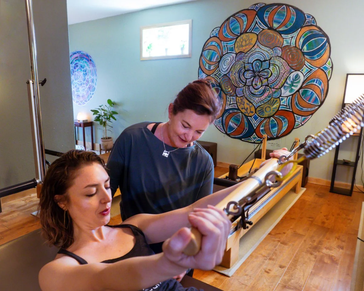 A woman practicing Pilates on a Reformer machine with a instructor assisting her, in a room with colorful wall art and a potted plant.