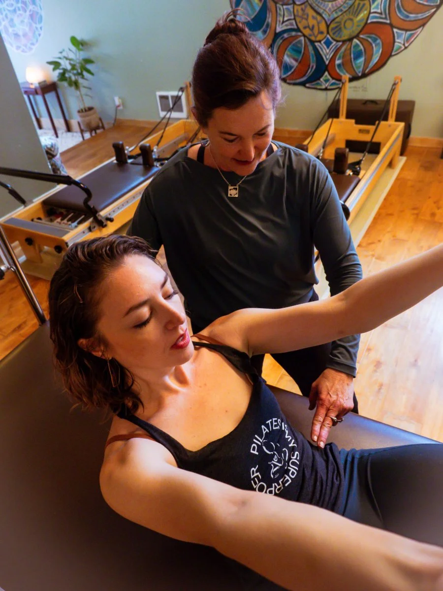 A woman performing a rollback with a rollback bar on a Pilates trapeze table with her arms extended; another woman standing beside her, assisting or guiding her in an exercise. The room has wooden floors, a plant, and colorful wall art.