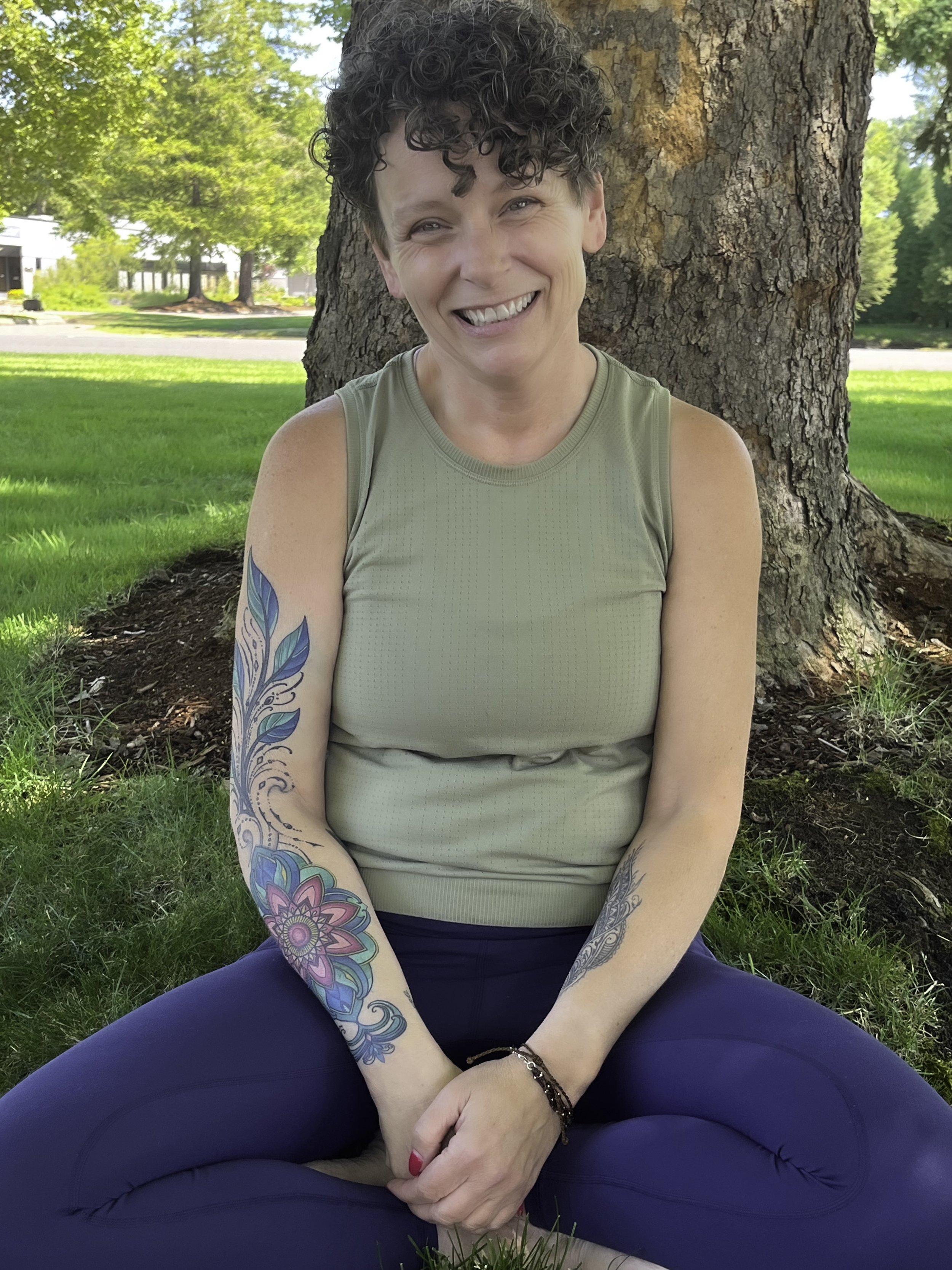 A woman with short curly hair and tattoos on her arms, sitting cross-legged on grass in front of a tree, smiling outdoors on a sunny day.
