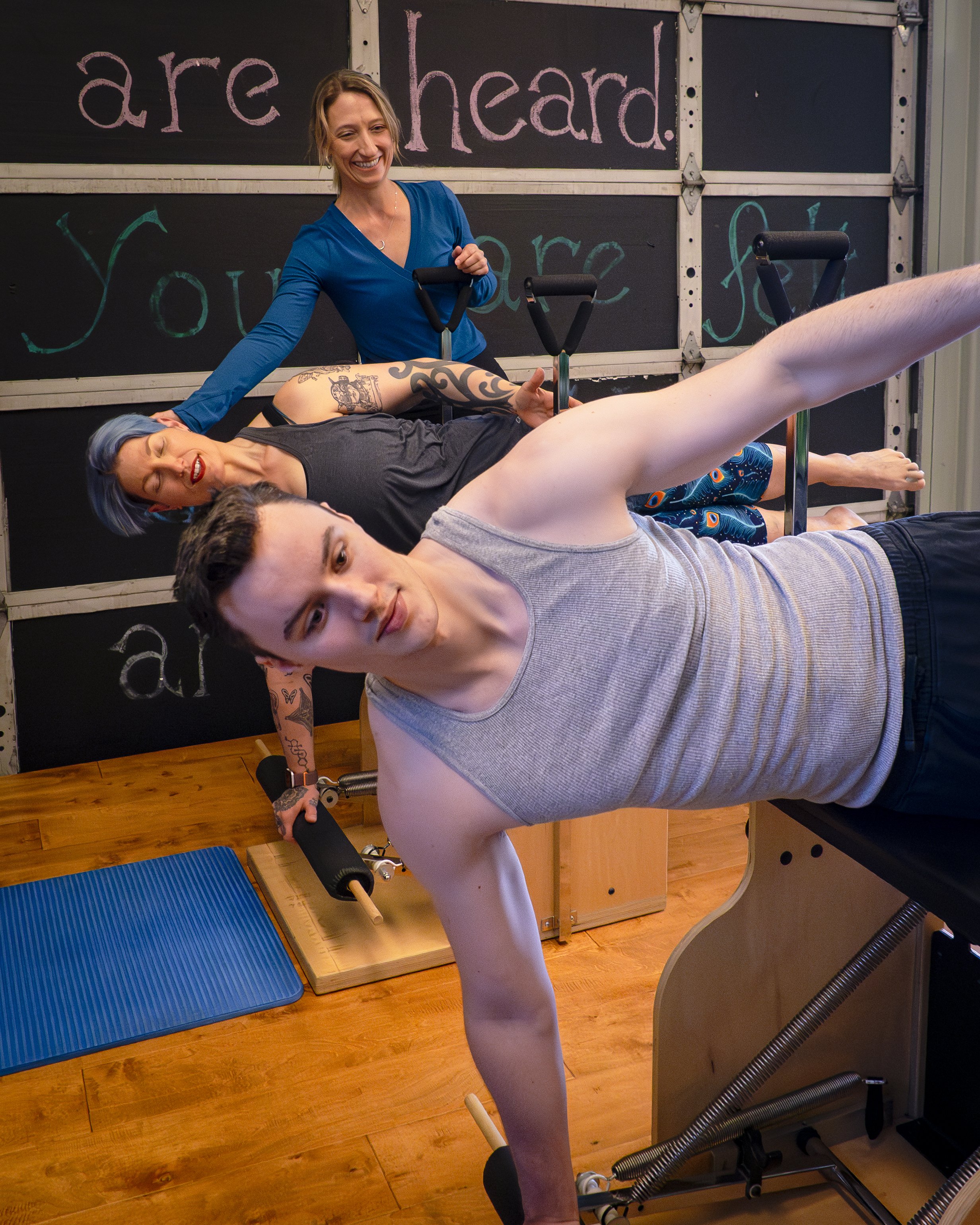 A woman  and man lying on their sides on Pilates wunda chairs in a Pilates studio with a chalkboard in the background that has inspirational words.