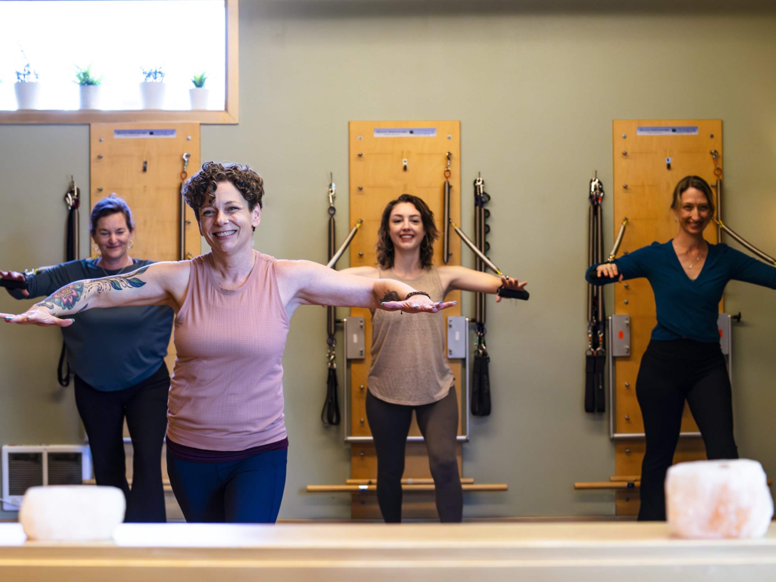 Four women participating in a Pilates springboard class, standing on Pilates mats in a studio with wooden springboards and a window with potted plants.