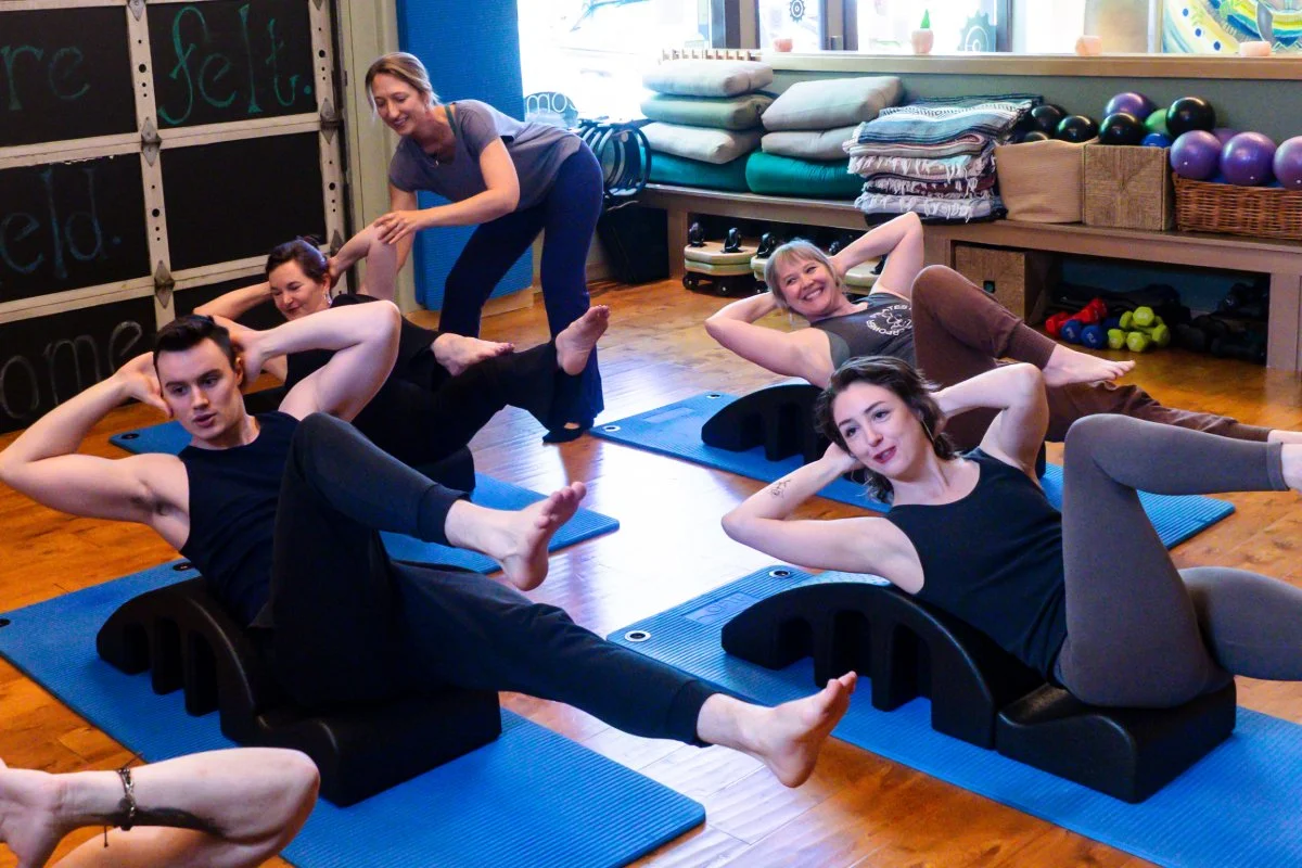 Group of five  participating in a Pilates class in astudio. They are lying on blue mats with Pilates equipment, and a woman instructor is assisting them. The studio has wooden floors, shelves with exercise balls, folded towels, and fitness accessories in the background.
