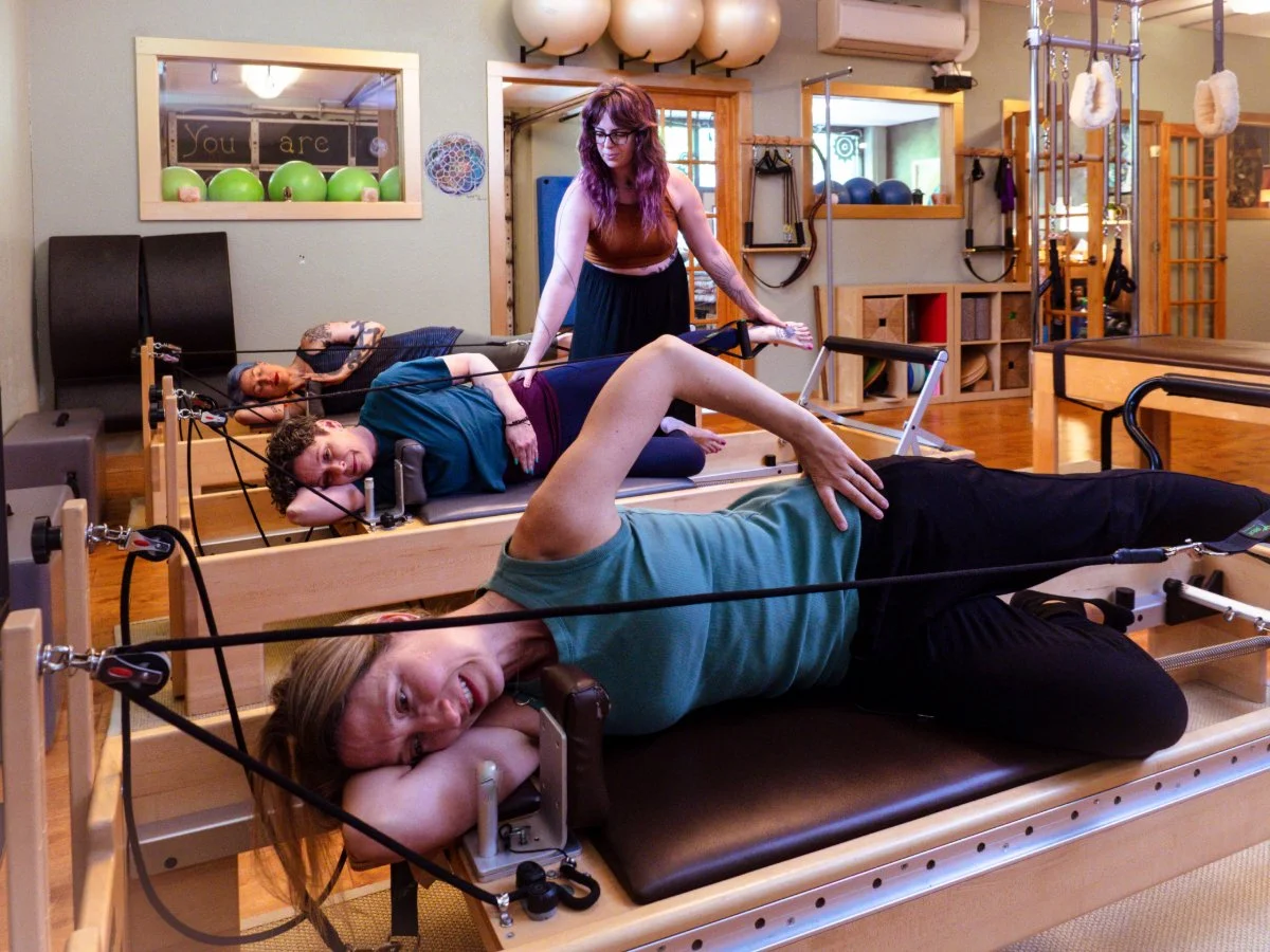 Women participating in a Pilates or physical therapy class using reformer machines in a gym studio.