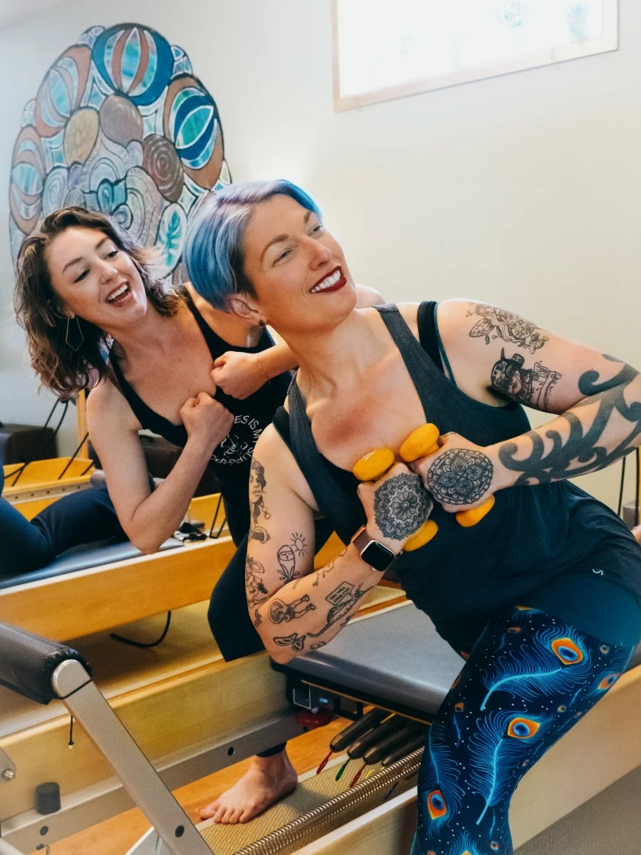 Two women exercising on a reformer Pilates machine, smiling and laughing, with one holding yellow weights and wearing colorful leggings, the other with decorated tattoos and wearing black clothes.