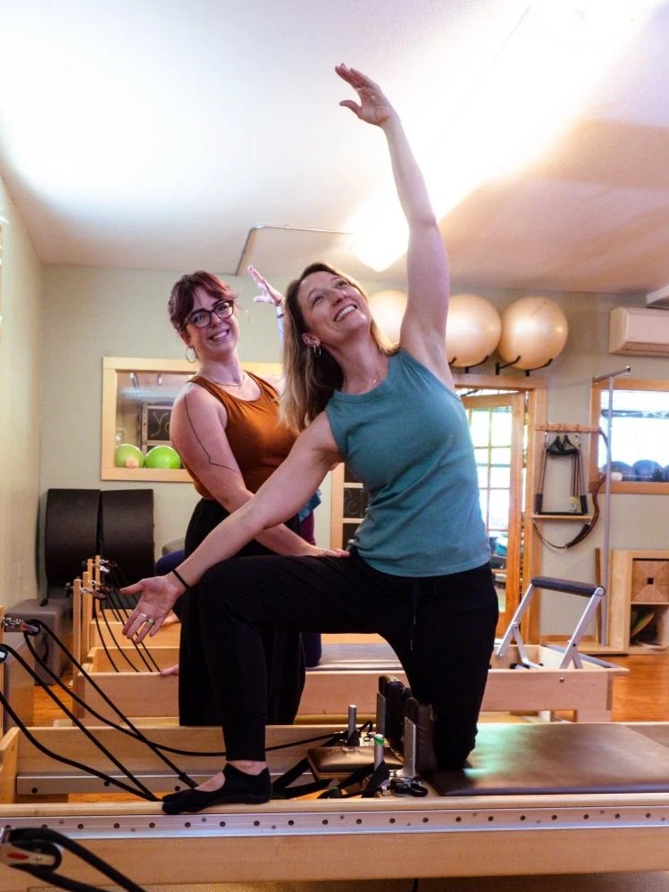 Two women with smiling expressions in a Pilates studio, one standing and the other kneeling on a reformer machine, performing Pilates exercises on a Pilates reformer.