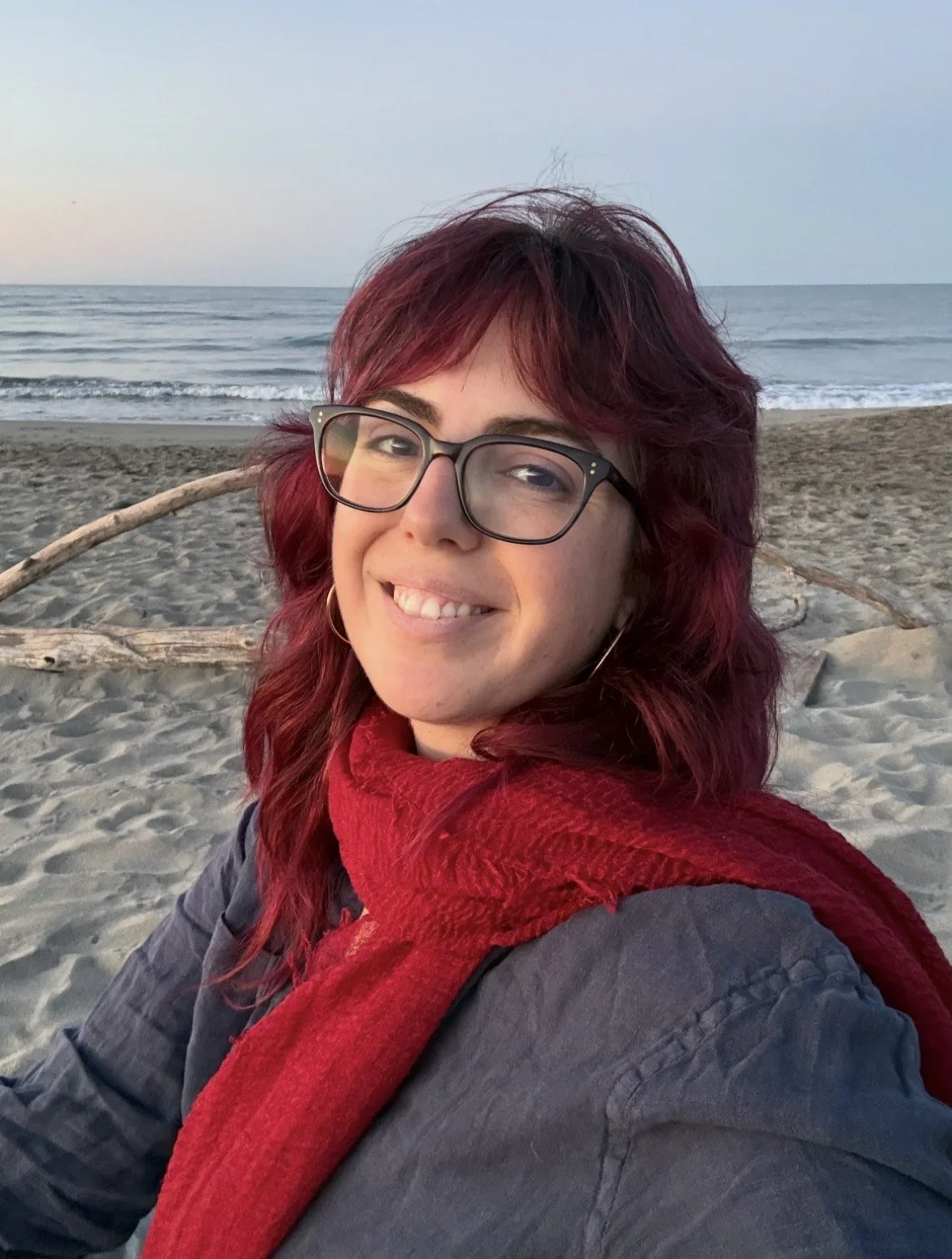 A woman with red hair and glasses smiling at the camera on a beach with sand, driftwood, ocean waves, and a pastel sky in the background.