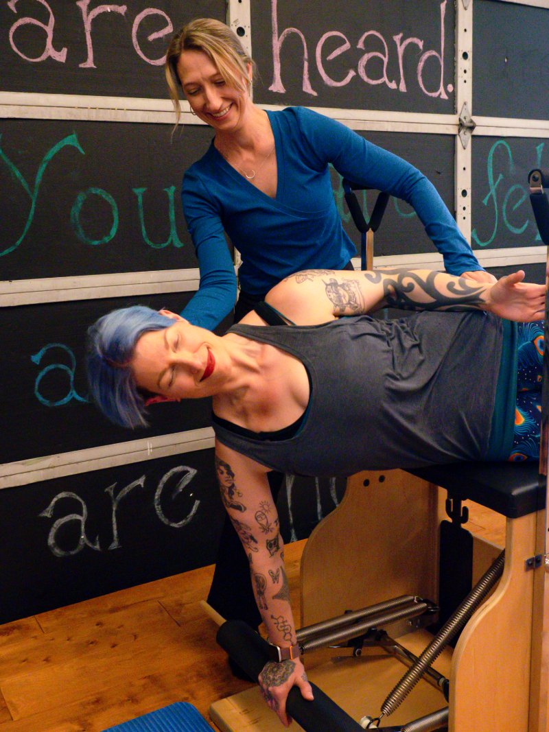 Two women in a Pilates studio. The woman with gray hair and tattoos is on a Pilates wunda chair while the other woman in a blue shirt assists her, smiling. There is a blackboard behind them with colorful writing.