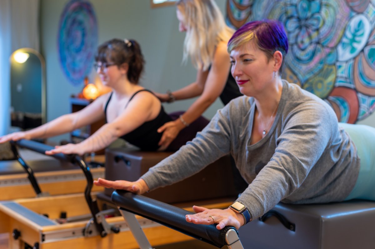 Women in a Pilates class doing stretching exercises on reformer machines, with colorful artwork on the wall in the background.