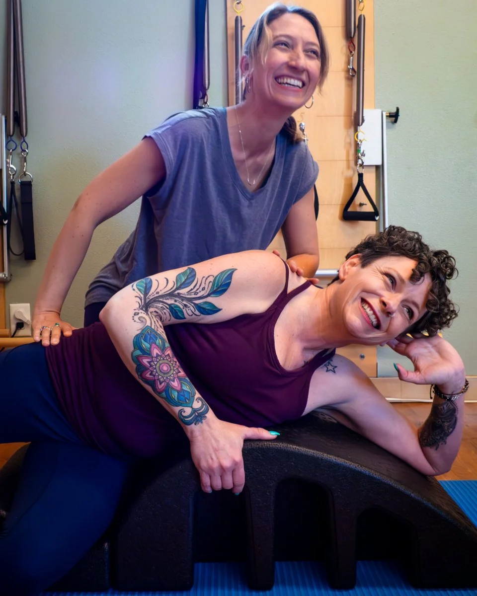 Two women smiling during a Pilates session, with one lying on a Pilates arc and the other leaning on her, in a Pilates studio with Pilates springboards in the background.