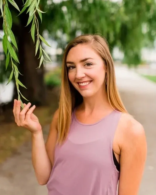 Smiling young woman with long blonde hair standing outdoors near a tree with green leaves