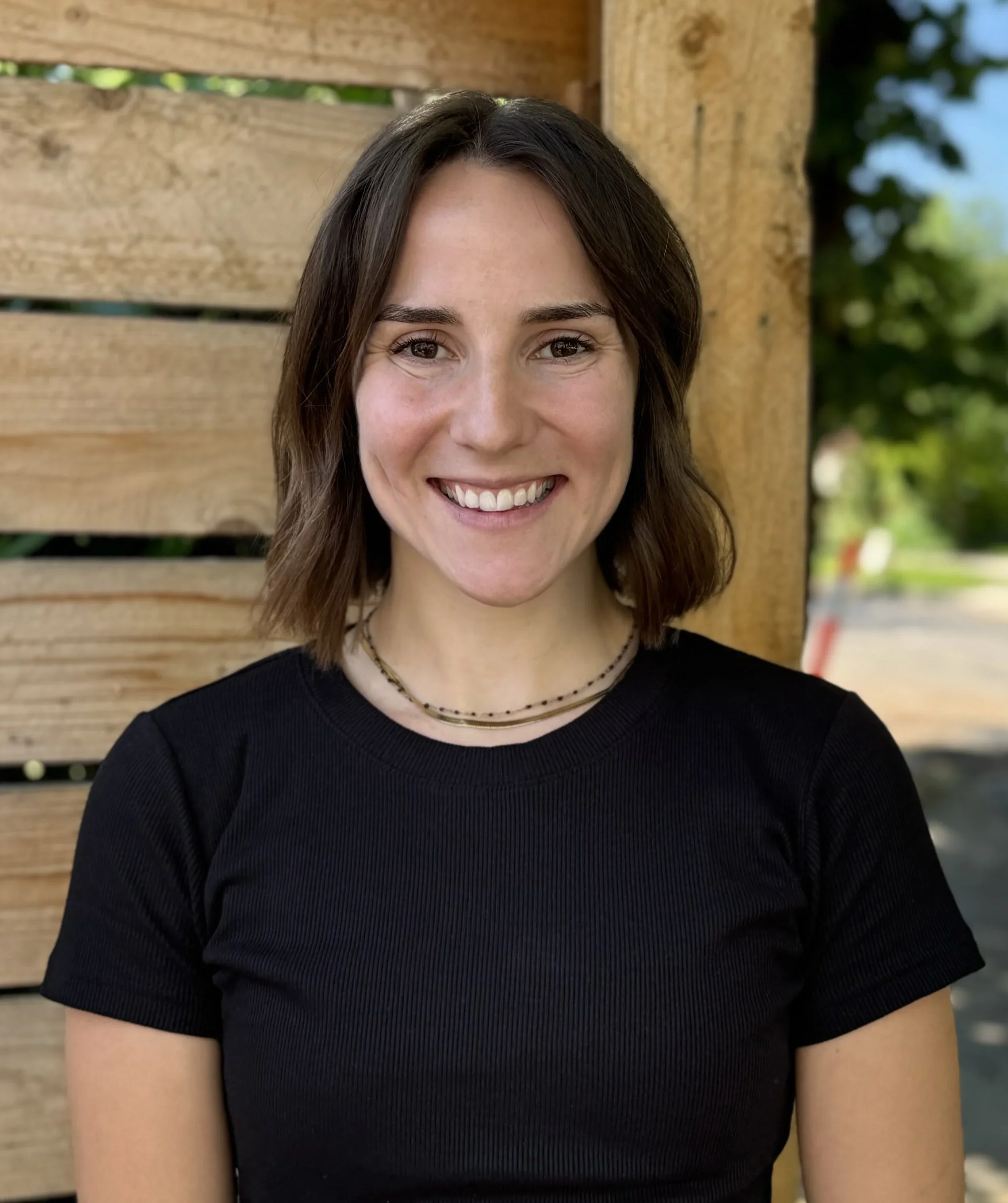 Close-up of a smiling woman with brown hair, wearing a black top and layered necklaces, standing outdoors near a wooden fence with leaves and trees in the background.