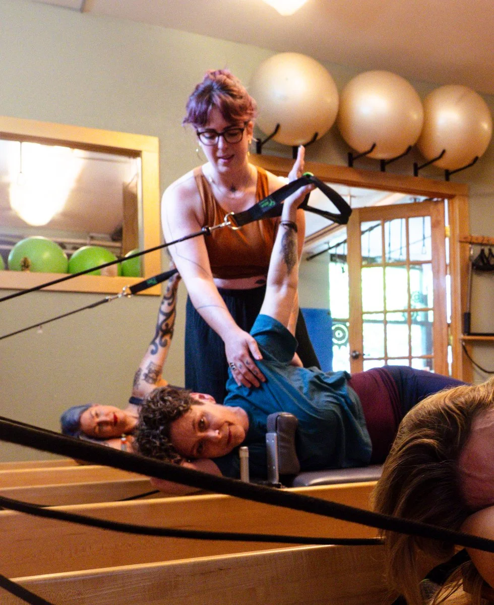 A woman assisting a woman in performing Pilates exercises on a Pilates reformer in a Pilates studio with exercise balls and mirrors.