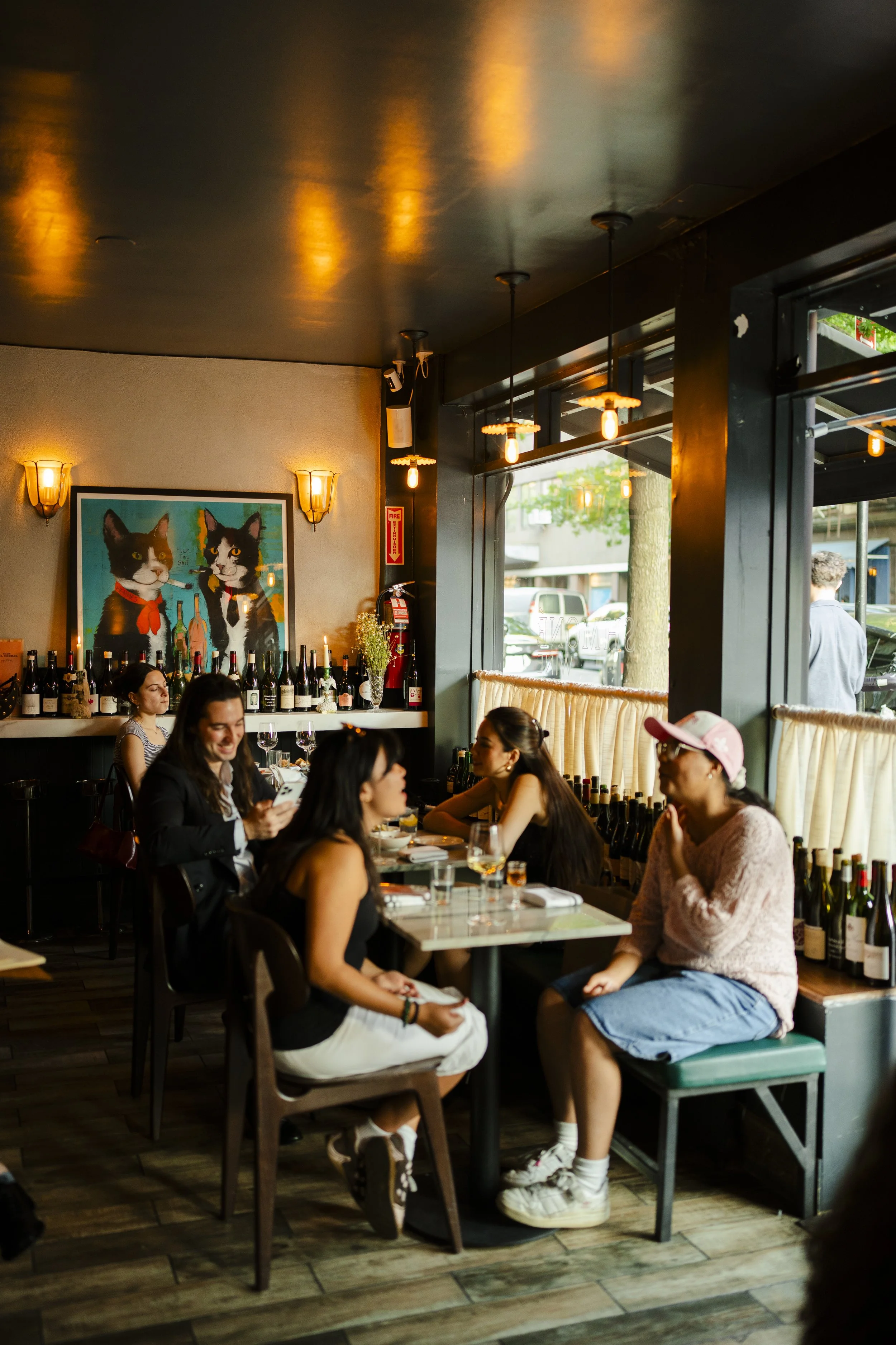 A group of women are sitting at a restaurant table with wine bottles, glasses, and plates, engaging in conversation. The restaurant has warm lighting, a large colorful painting of two cats on the wall, and large windows letting in natural light.