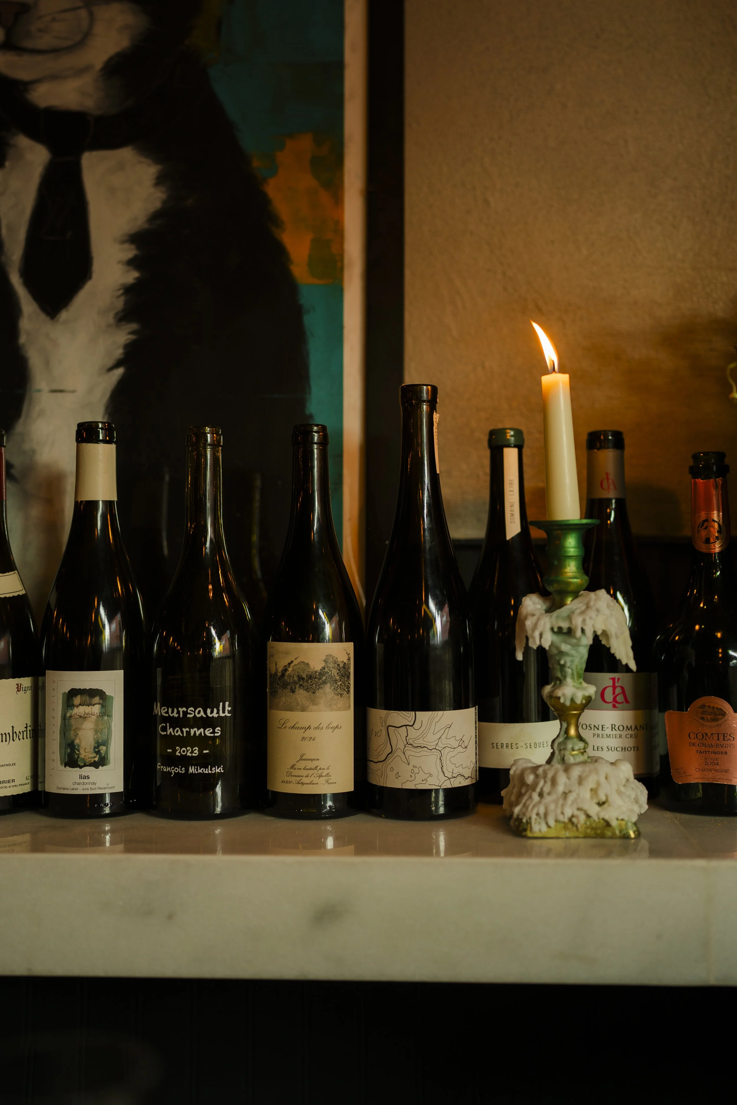 A row of wine bottles on a shelf with a lit candle in a green and gold candle holder in front of them, and a colorful painting in the background.