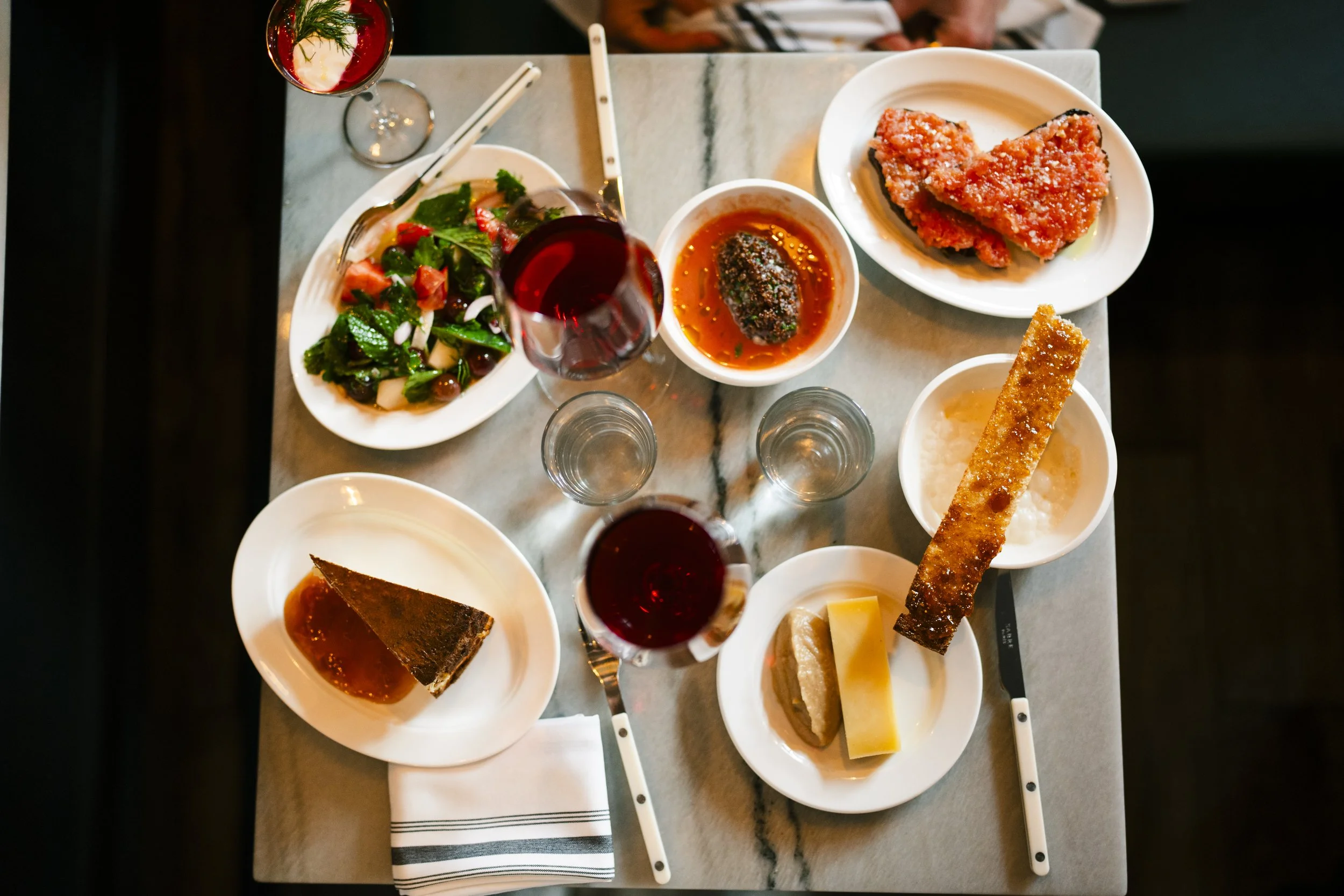 A variety of dishes on a marble table, including salads, meats, desserts, and glasses of red wine.